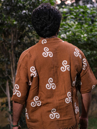 A person with curly hair stands facing away in a Spiral Kurta, featuring blockprinted tribal spiral designs on a brown fabric. They are outdoors, surrounded by lush green foliage.