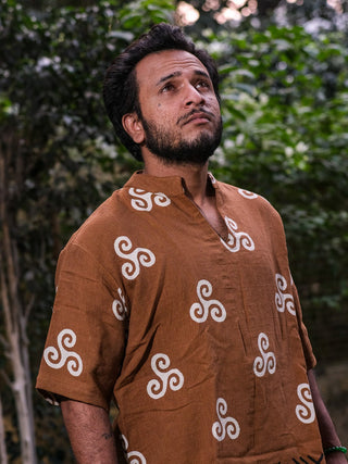 A man with short dark hair and a beard, wearing a brown Spiral Kurta, gazes up thoughtfully while standing outdoors in front of green foliage.