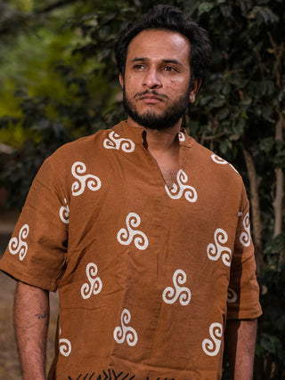 A man with a beard and short hair wears the Spiral Kurta, a brown crepe shirt with white tribal spiral designs, standing outdoors before green foliage and looking to the side with a serious expression.