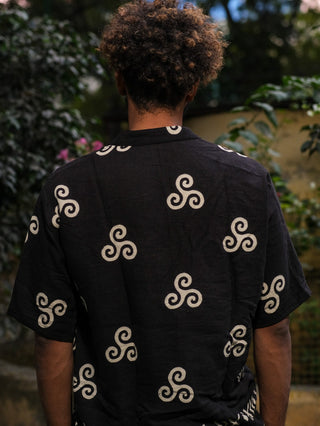 Seen from behind, a person with curly hair wears a black Spiral Kurta displaying a white triskelion design with boho flair. The lush greenery and blurred outdoor backdrop contribute to the serene ambiance.