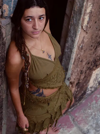 A woman with long brown hair stands in a rustic stone doorway, wearing an olive green Titania Crop Top. She has tattoos on her chest and side, and wears a round pendant necklace, gazing up toward the camera.