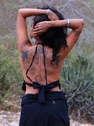 A person with dark curly hair stands outdoors, back to the camera and arms raised. Intricate black tattoos cover their back, and they wear the Titania Crop Top with black textured pants.