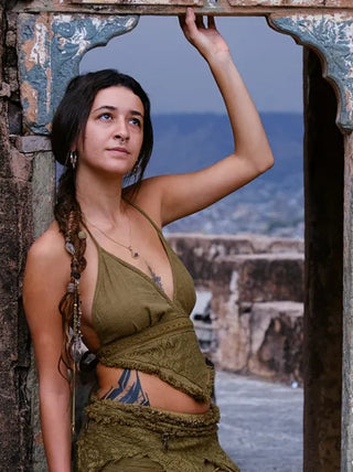 A woman with long braided hair and tattoos stands in an arched stone doorway, wearing the Titania Crop Top. She gazes thoughtfully upward, with mountains and ancient ruins visible behind her.