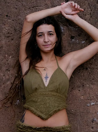 A woman with long brown hair poses against a textured brown wall, wearing an olive green Titania Crop Top and necklace, her arm raised above her head and a gentle expression on her face.